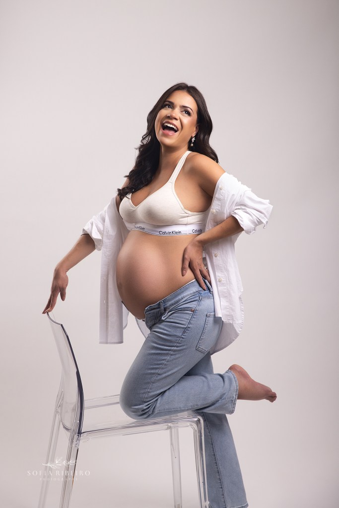 a joyful portrait as mom laughs in a white shirt and jeans, belly exposed during a photo session with a cedar grove nj maternity photographer