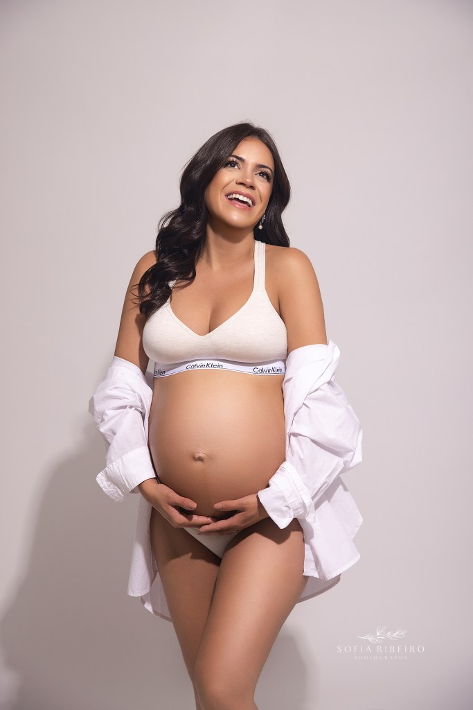 mom beams with joy in a white shirt and underwear combo, cradling her baby bump for maternity portraits in studio