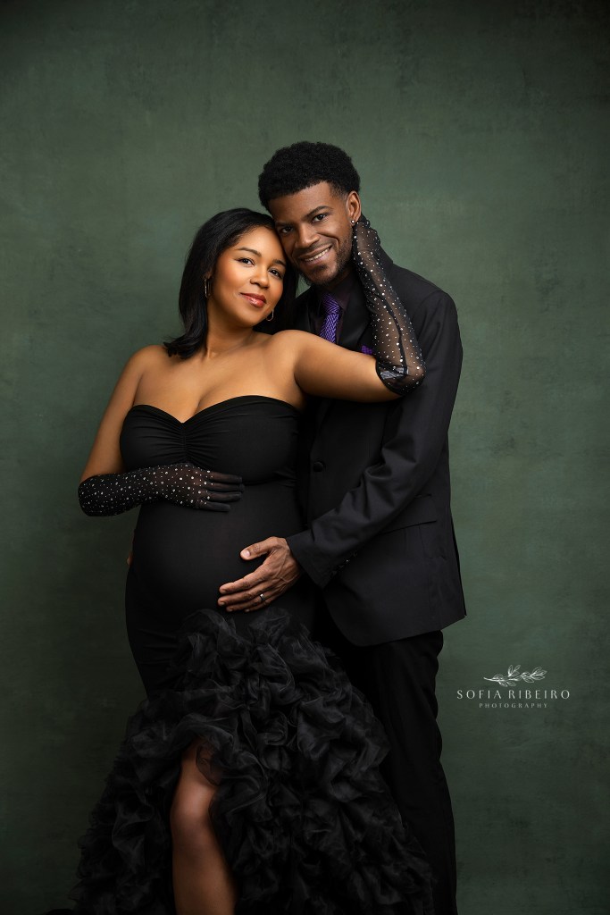 portrait of mom and dad to be against a green painted backdrop, in black wardrobe cradling the belly and smiling to camera, shot by a luxury nj maternity photographer
