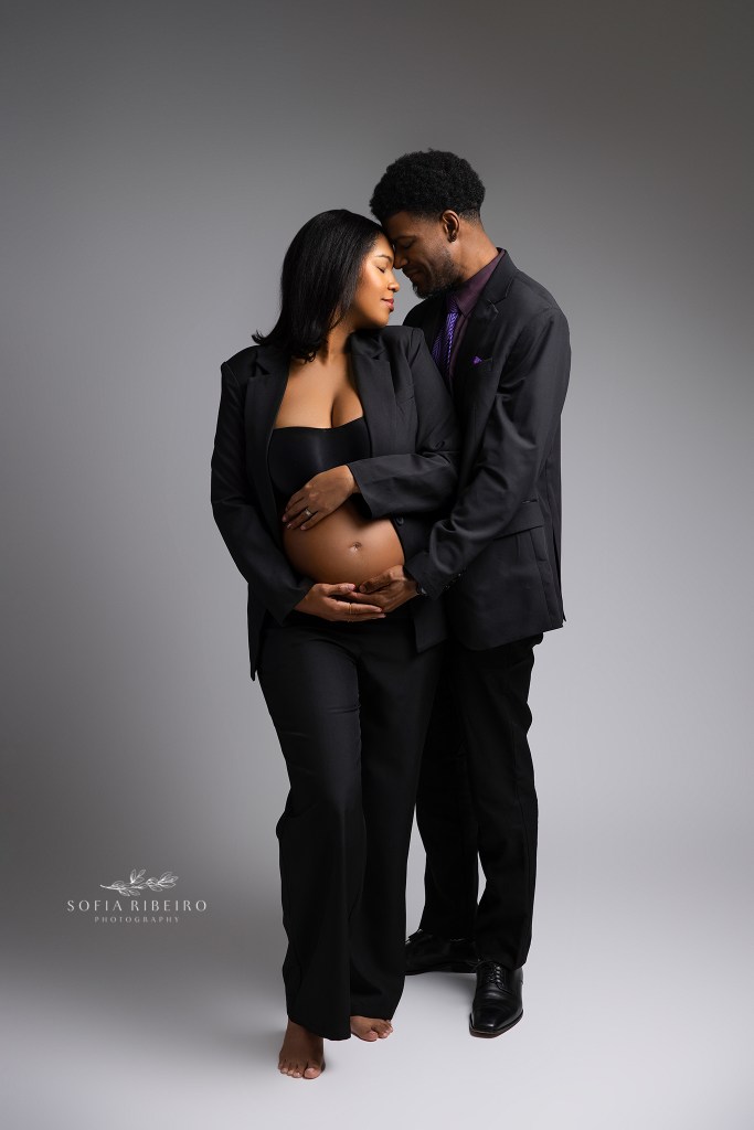 a power suit moment for expectant mom and dad, cradling the baby bump in matching black pantsuits against an ombre grey backdrop. shot by high end nj maternity photography studio in dunellen nj