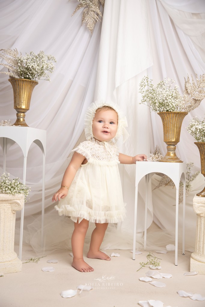 a baby girl in a sweet white romper stands up next to some gold planters and flowers for a mini mommy and me session