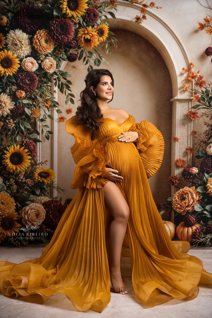 a pleated golden gown showcases mom's baby bump against a floral backdrop for maternity photos