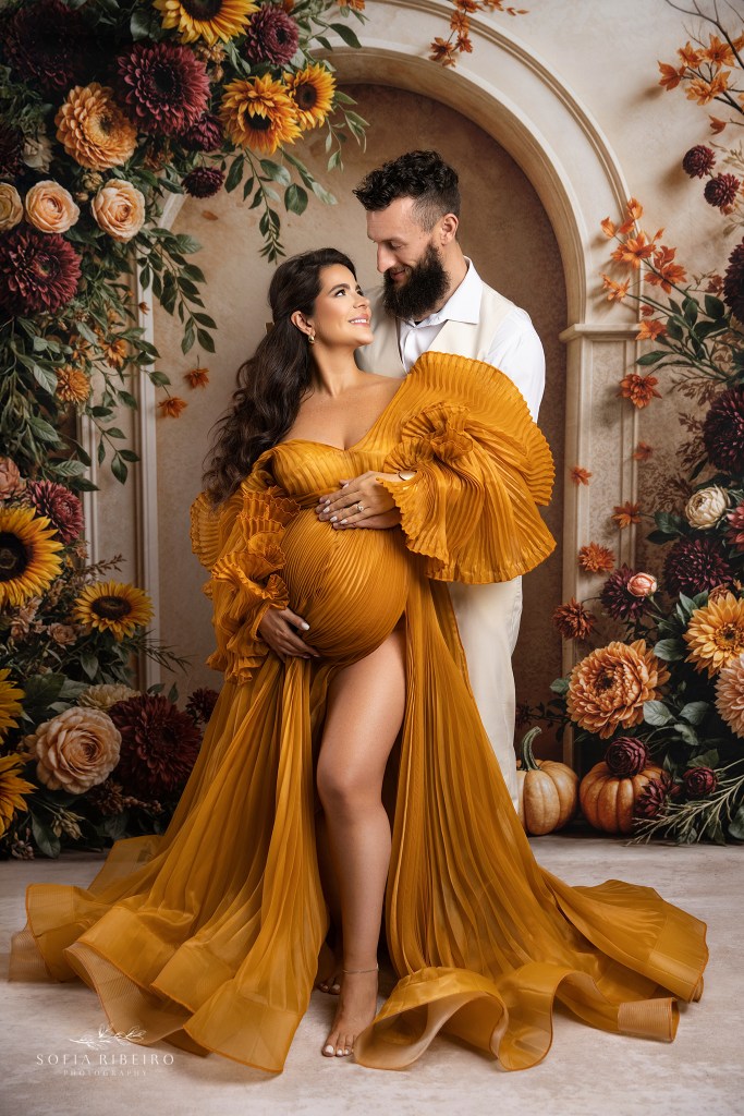 mom and dad are set against a floral backdrop, in fall hues, golden dress cradled against the baby bump during a pregnancy photoshoot in south jersey