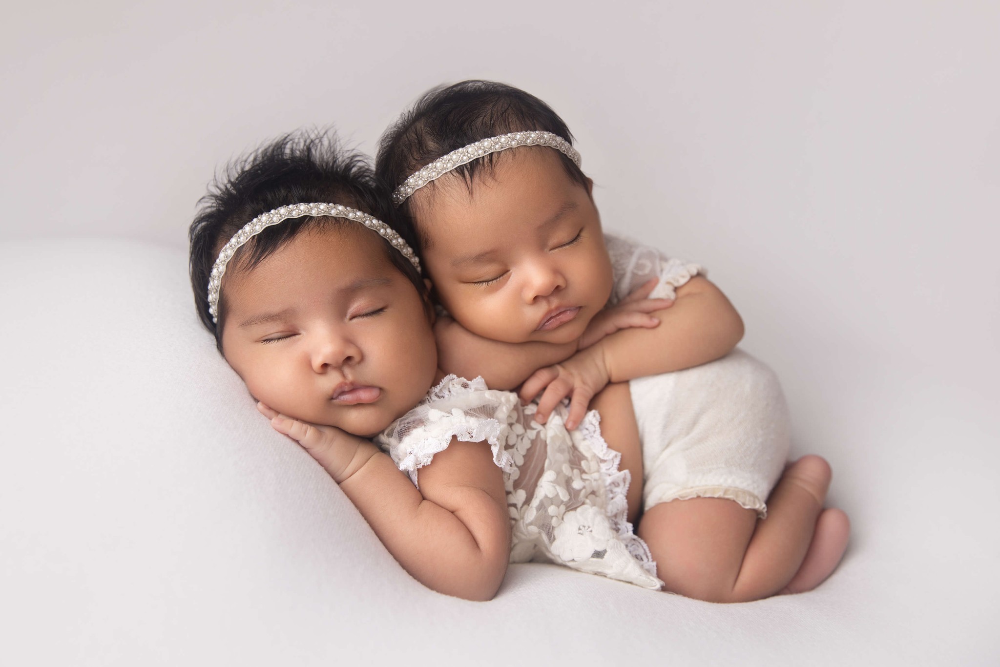 Newborn baby twin sisters in white lace shirts sleep on top of each other in a studio before visiting daycares in westfield nj