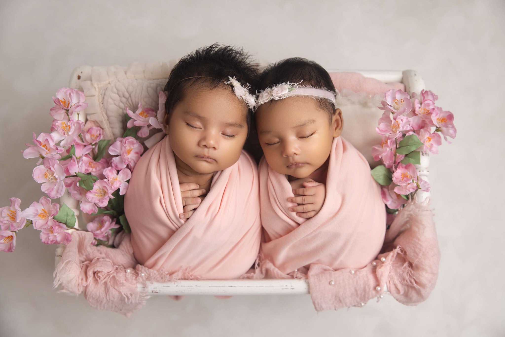 Twin newborn baby sisters sleep in a tiny wooden crib with flowers and matching pink swaddles before finding daycares in westfield nj