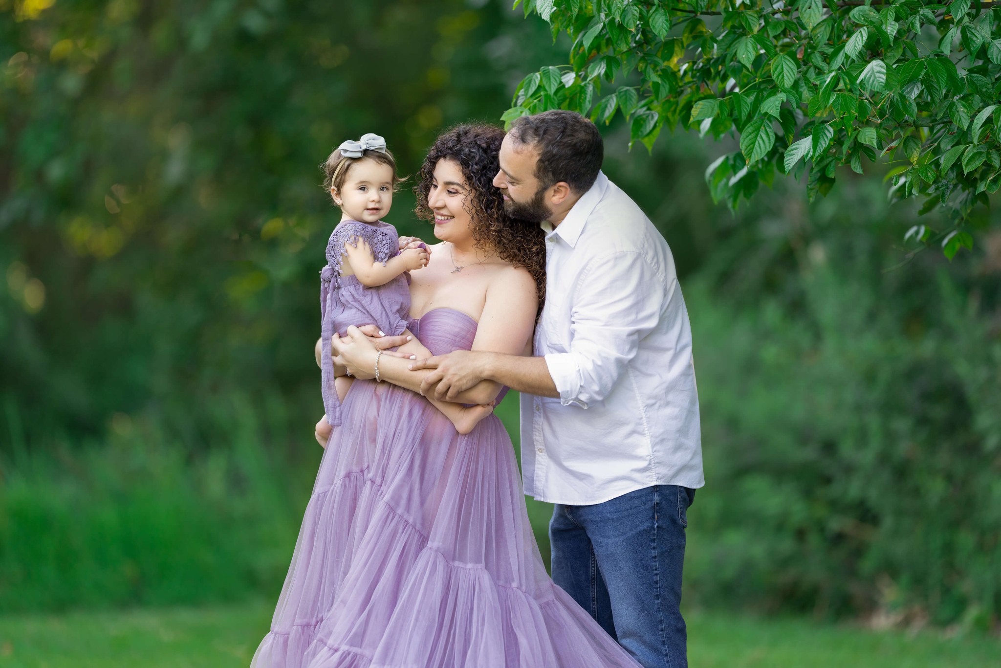 Happy mom and dad in a white shirt and purple gown play with their toddler daughter in matching purple in a park after exploring preschools in westfield, NJ