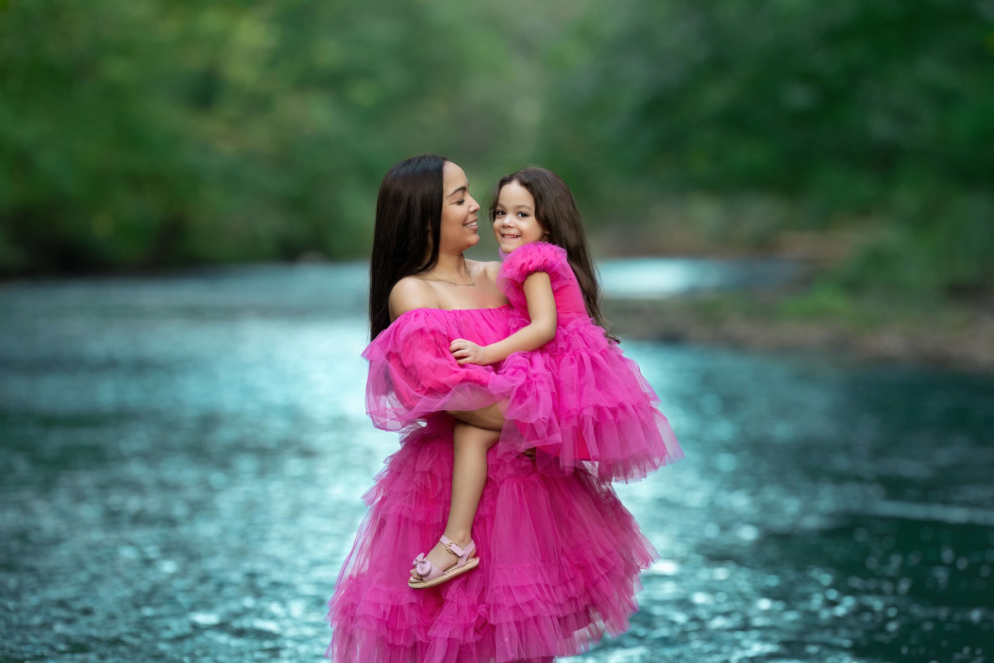 A smiling mother stands on the edge of a river holding her toddler daughter as they both wear matching pink tulle gowns before some gymnastics in union county, NJ