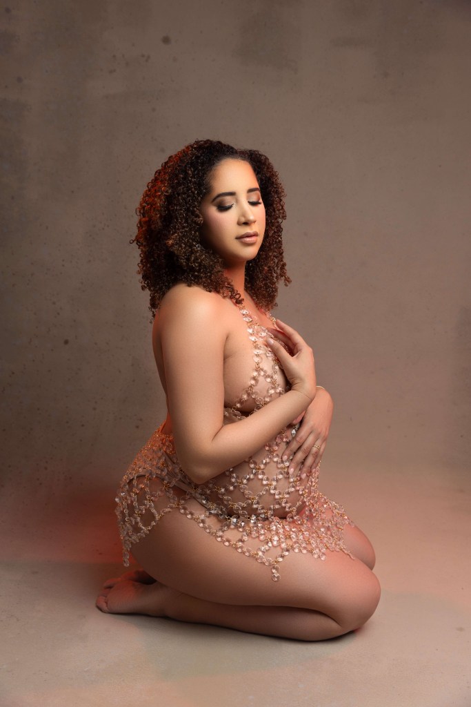 A mother to be kneels in a rhinestone bodysuit on the floor of a studio with eyes closed