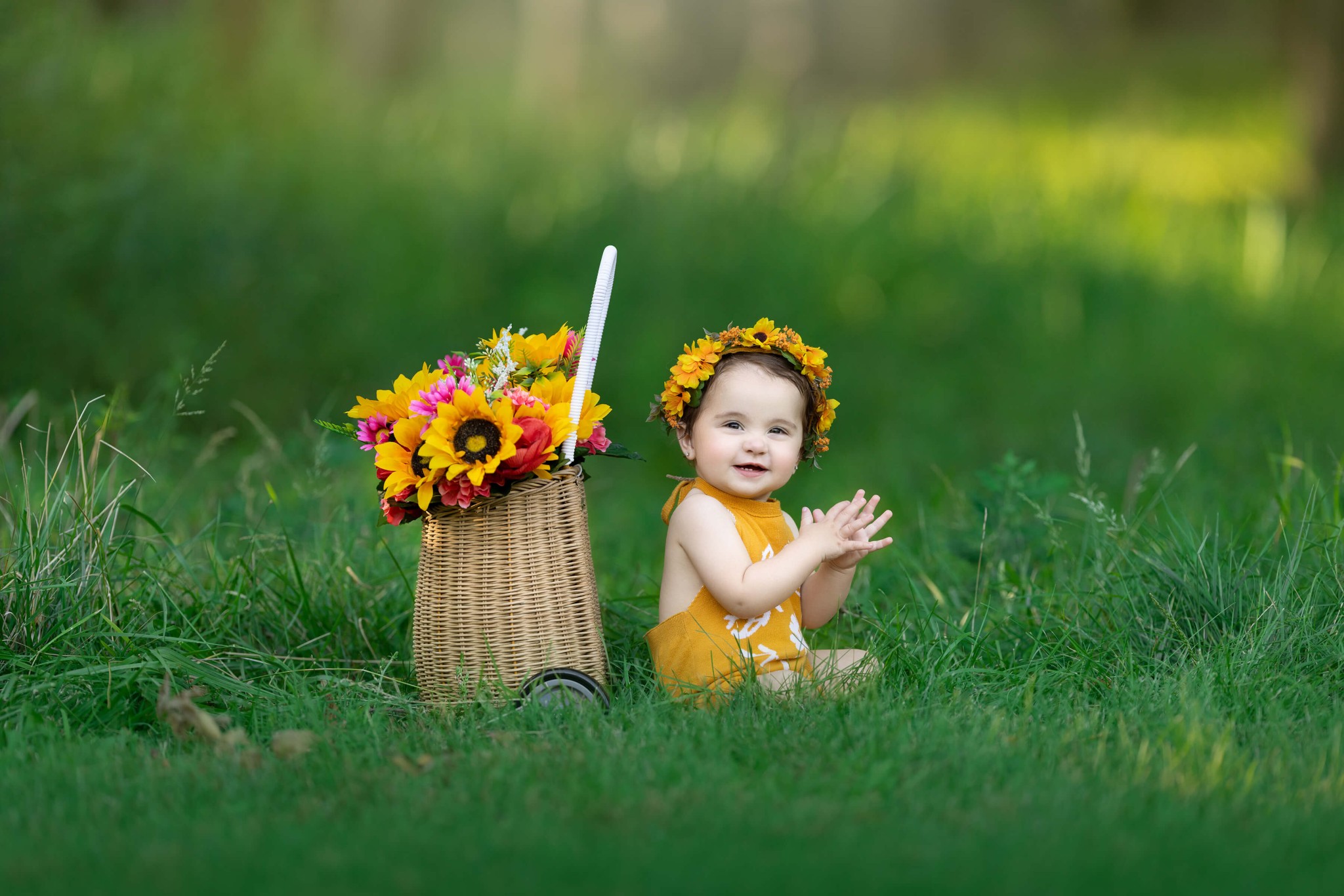 A happy baby girl in a yellow dress and flower headband sits in the grass with a woven flower cart after visiting pediatric dentists in westfield, NJ
