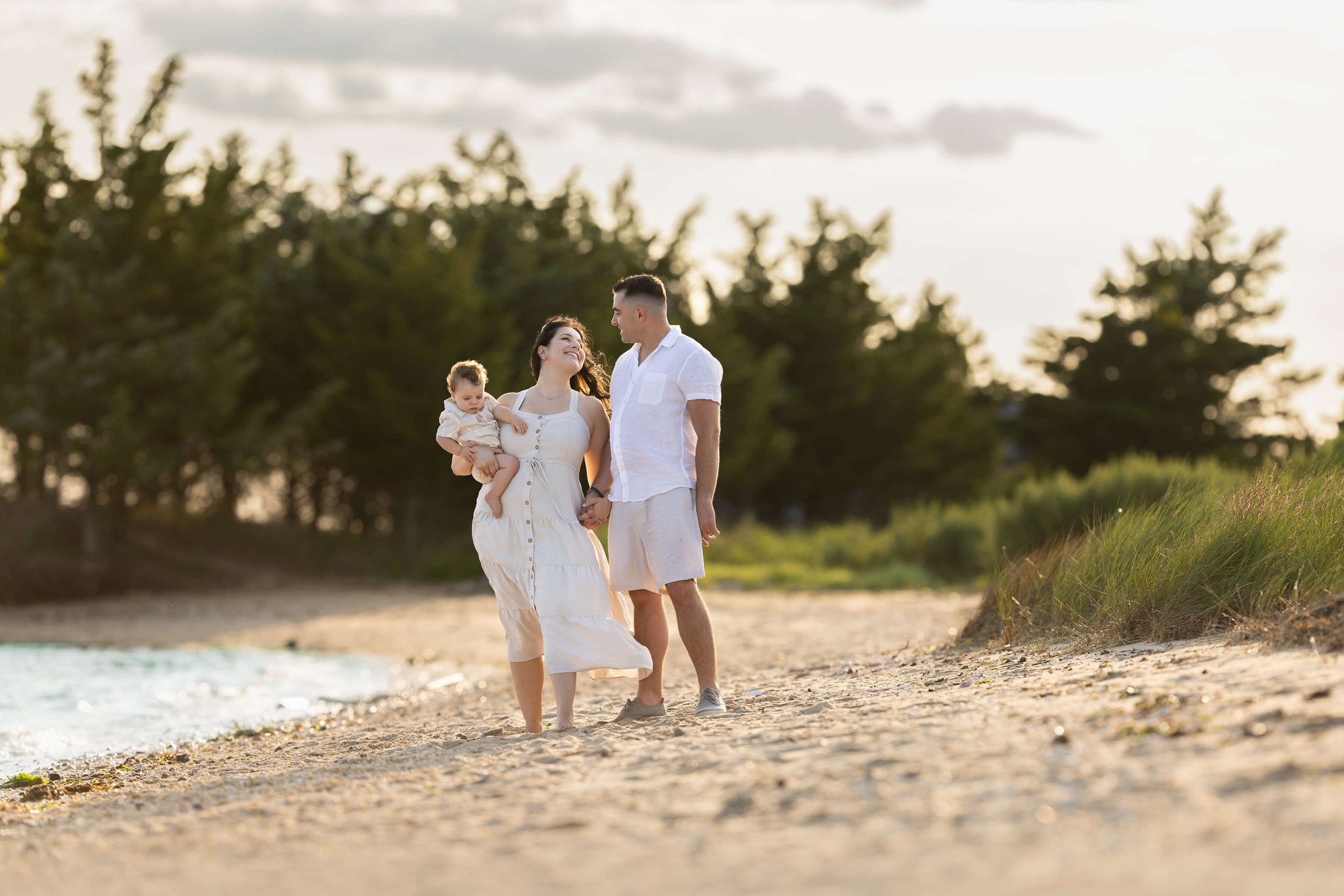 A mom and dad walk on a beach at sunset in white while mom holds their toddler on her hip