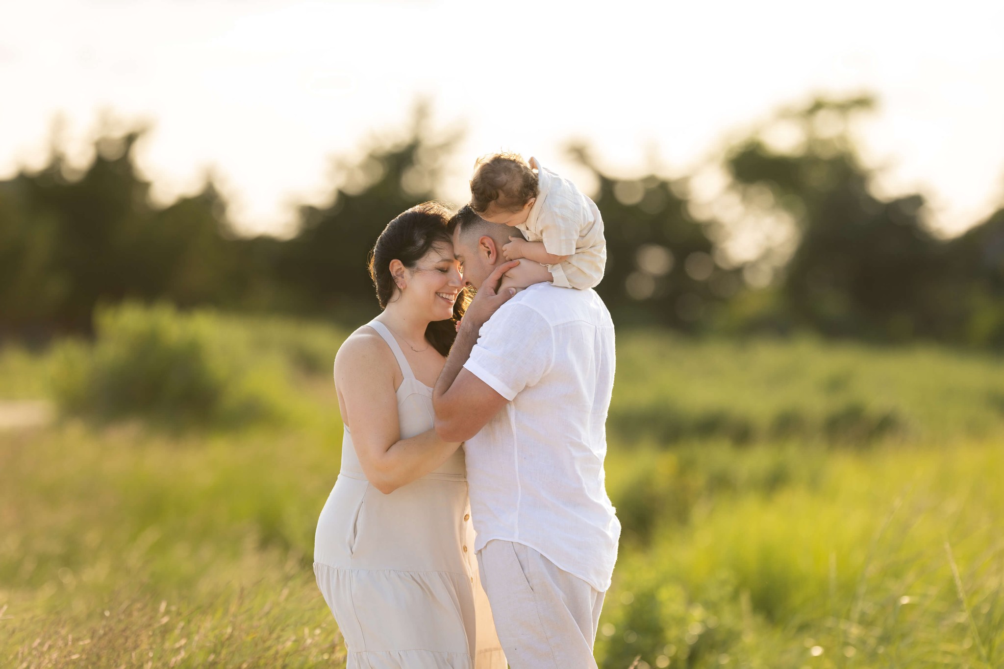 Happy mom and dad snuggle touching foreheads on a beach in white outfits with a toddler on dad's shoulders at sunset after visiting playgrounds in union county