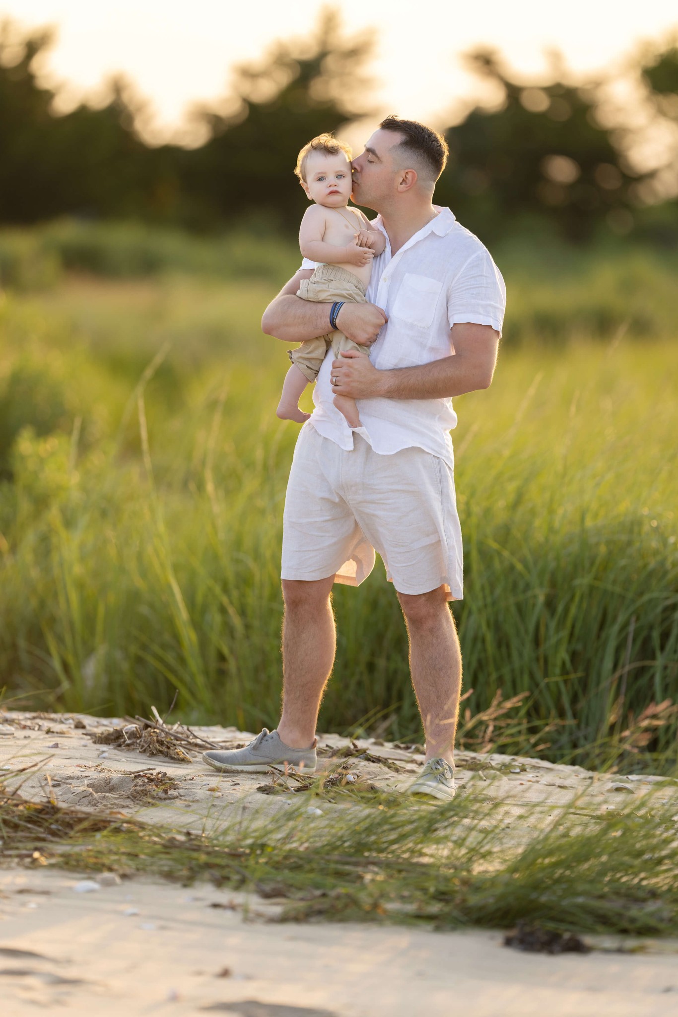 A dad in a white shirt kisses the cheek of his toddler son in his arms on a beach dune at sunset after visiting playgrounds in union county