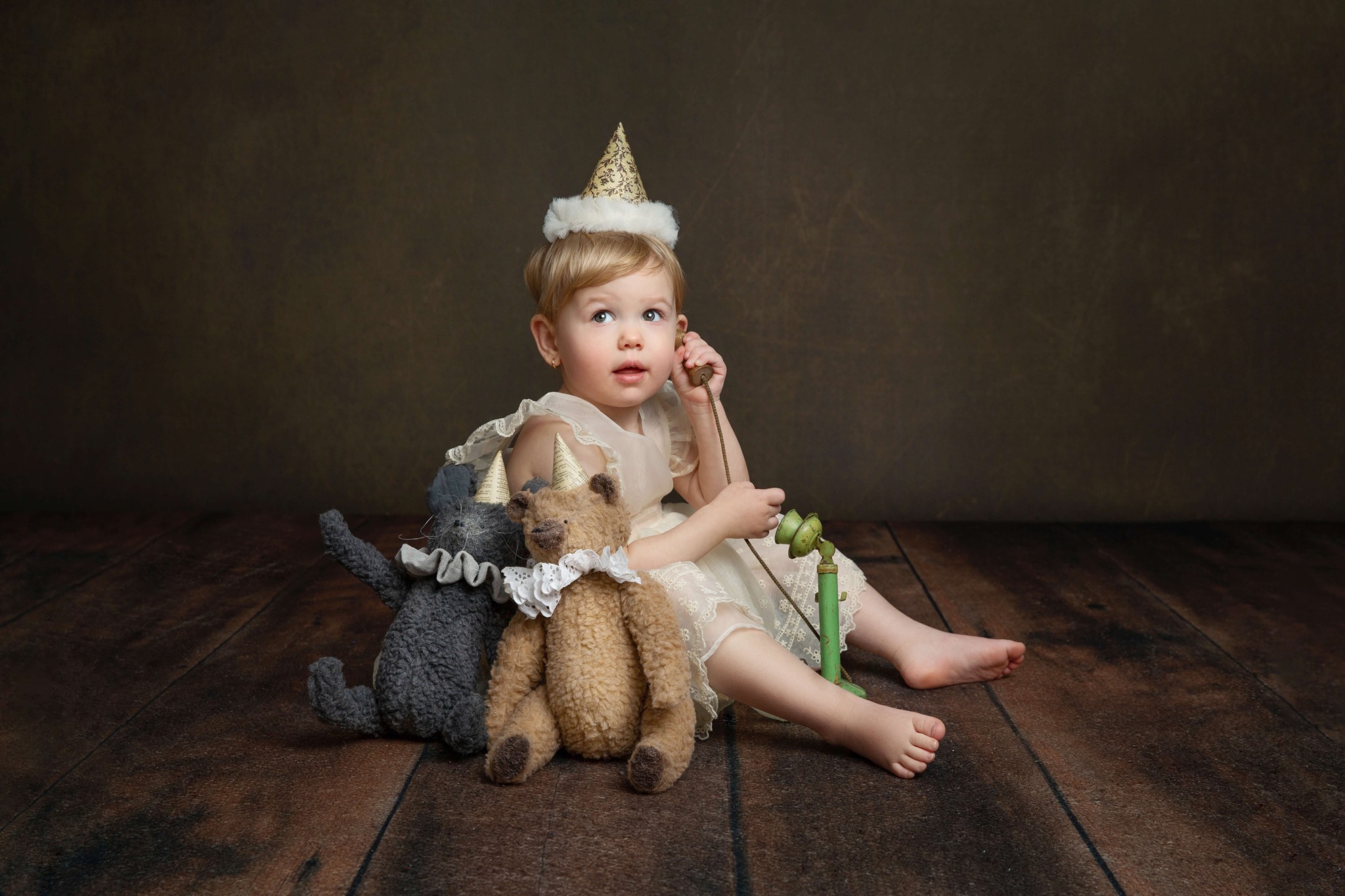 A toddler girl in a white dress and birthday cap sits on a toy vintage telephone and two teddy bears