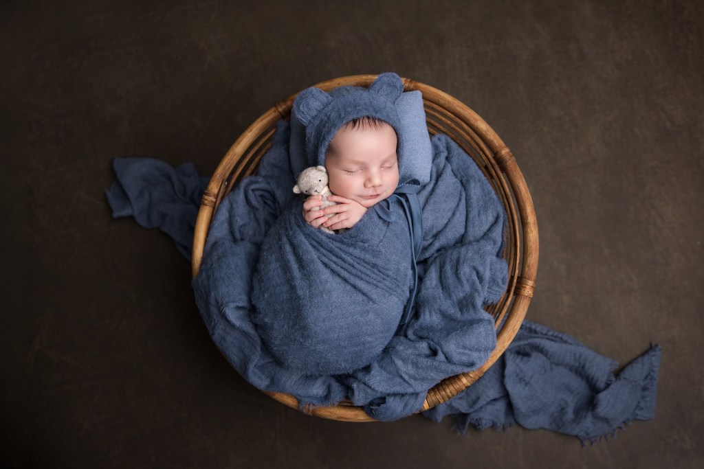 baby boy is posed in a circular basket with blue wrapped fabrics and a teddy bear in a nj newborn photo studio