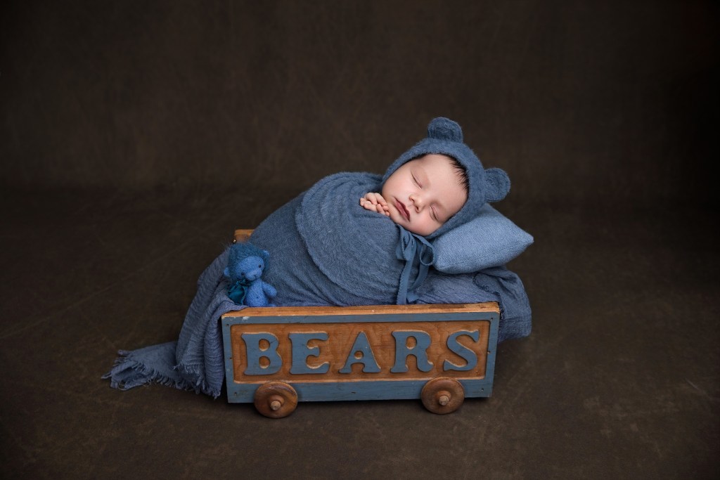 a newborn photographer poses a baby boy in a bear hat and rolling cart