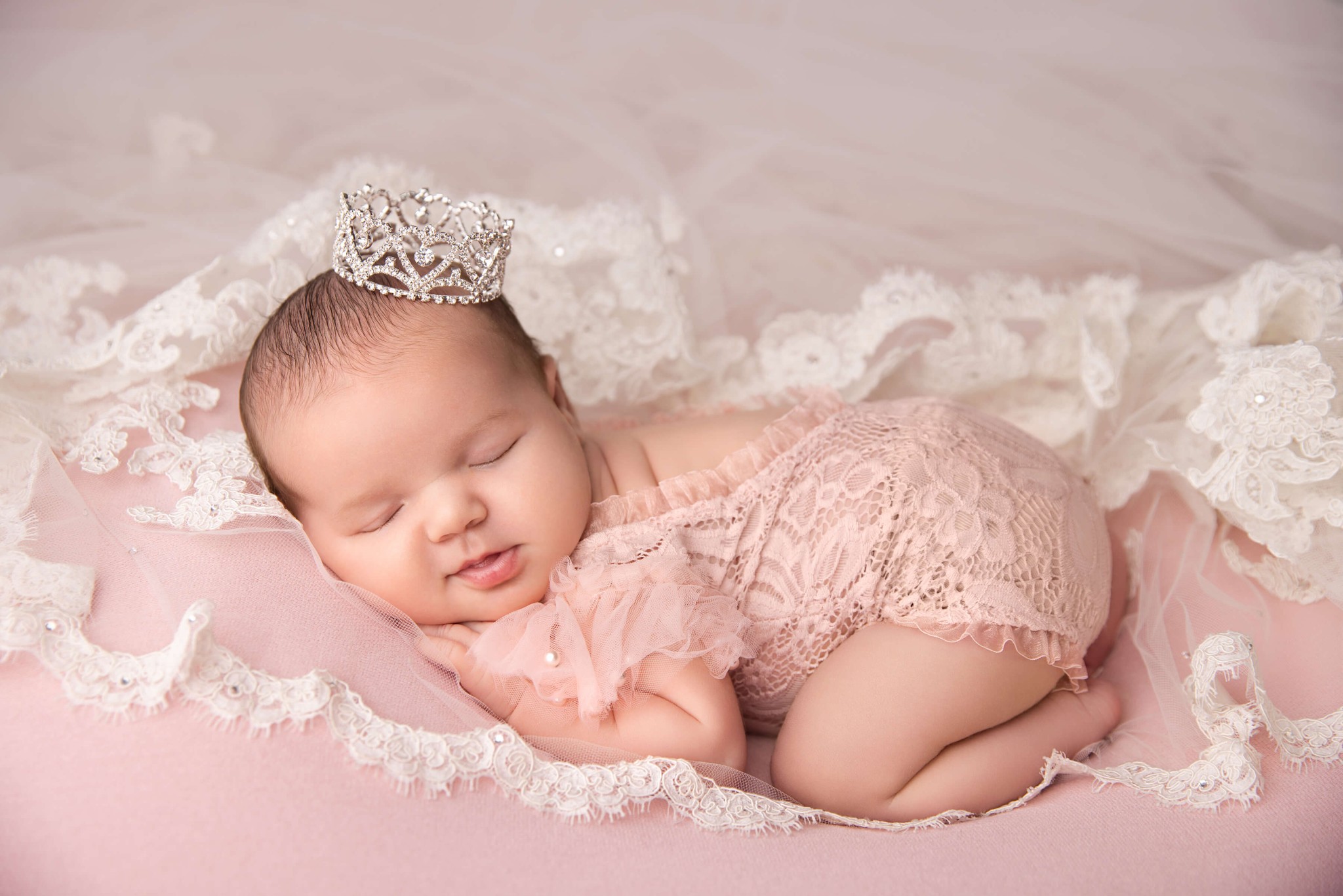 A newborn girl in a pink lace onesie sleeps on her tummy on a lace veil and pink bed after finding baby sleep consultants in nj