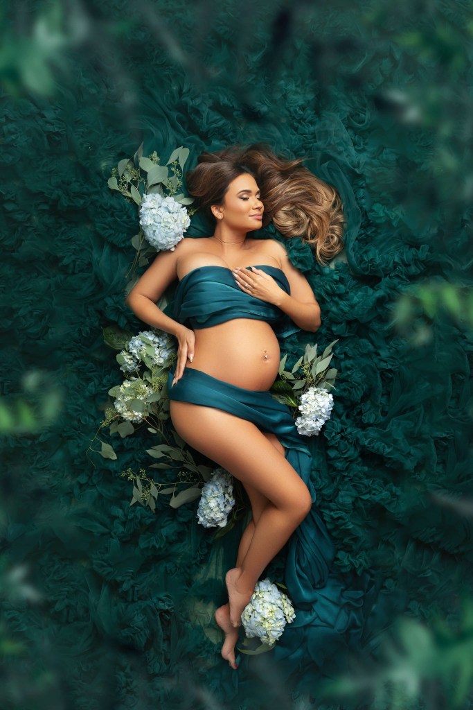 A pregnant woman lays on a bed of teal tule with a matching silk wrapped around her after enjoying fertility acupuncture in nj