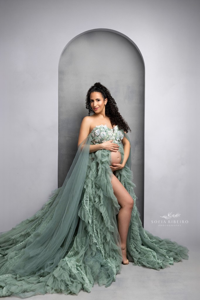 mom to be dressed in a sage green tulle and lace gown against a grey backdrop