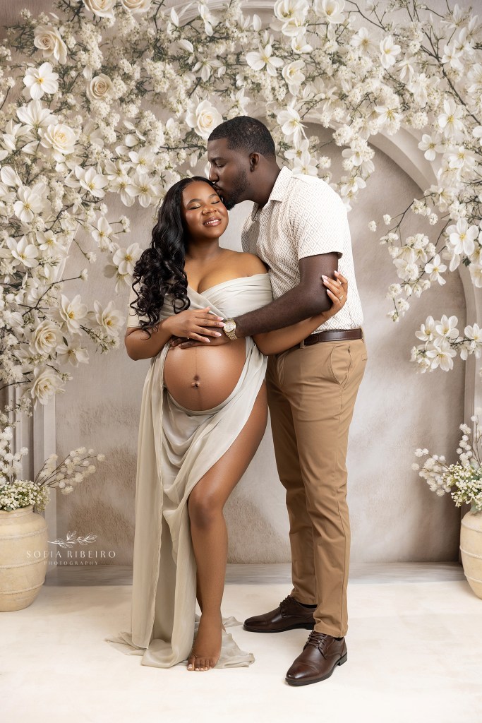 mom and dad share a sweet moment together against a white blossom backdrop, cradling the baby bump for maternity photos