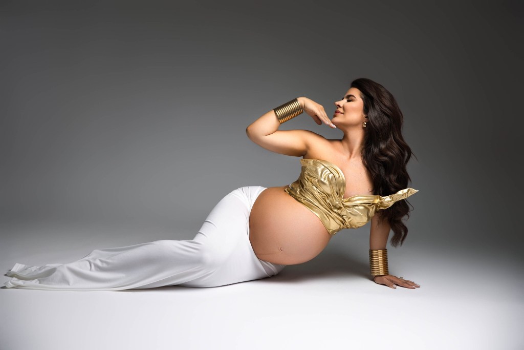 A smiling pregnant woman lays across the floor of a studio with a hand under her chin for a nj maternity photographer