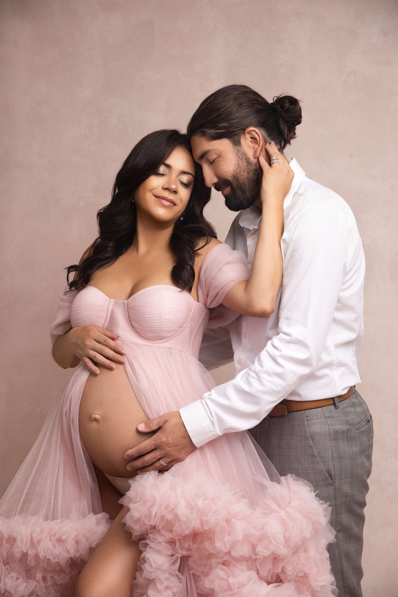 A happy pregnant couple snuggle in a studio as mom stands in pink tule maternity gown after visiting obgyns in morristown, NJ