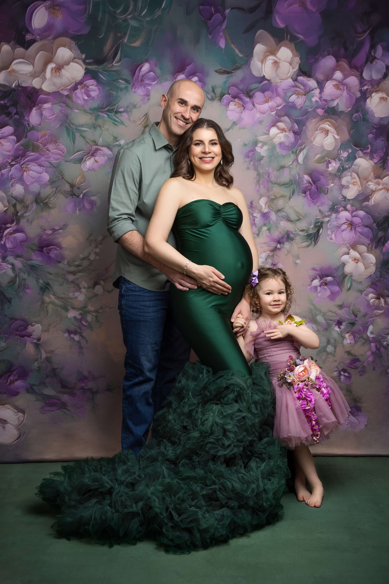 A happy mother to be in a green maternity gown stands in a studio with her husband and toddler daughter after some prenatal yoga in north jersey