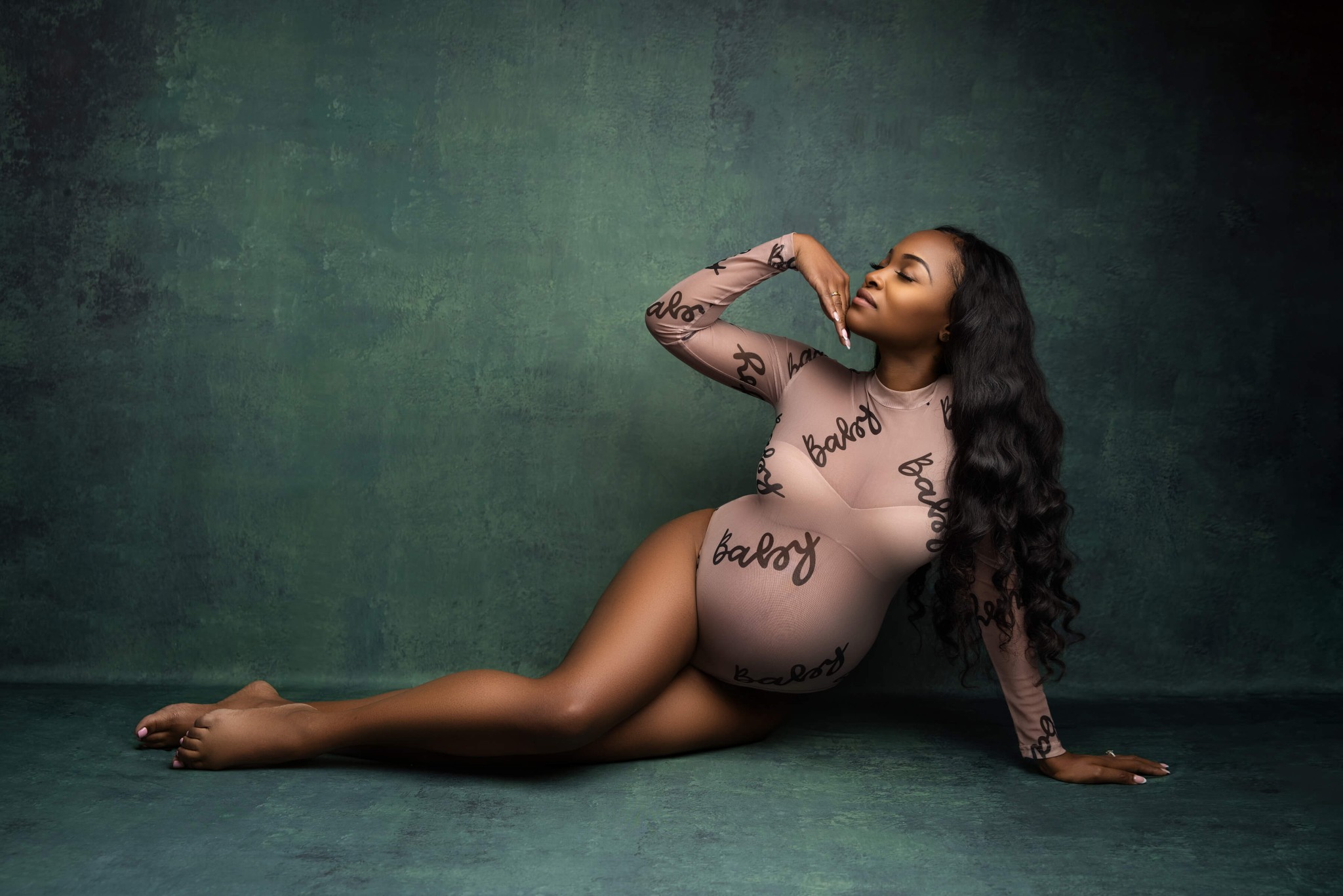 A woman in a pink bodysuit sits across to floor in a studio with fingers under her chin leaning on her other hand