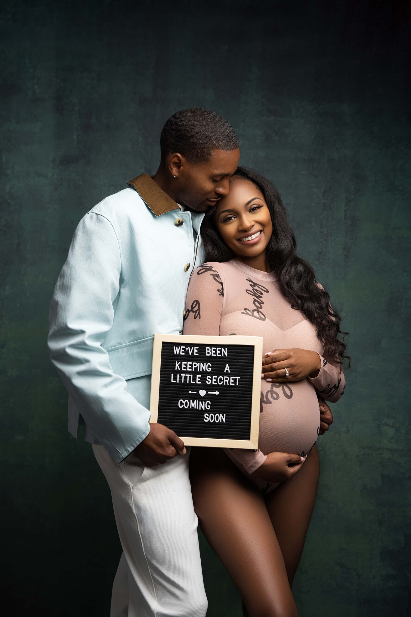 A happy couple snuggle while holding a baby announcement sign in a studio after enjoying prenatal yoga in union county nj