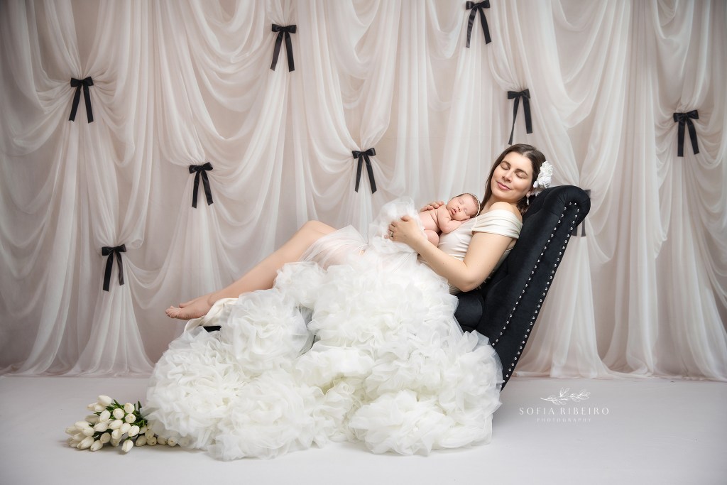 mom and baby pose elegantly on a black recliner in a white gown for newborn photos