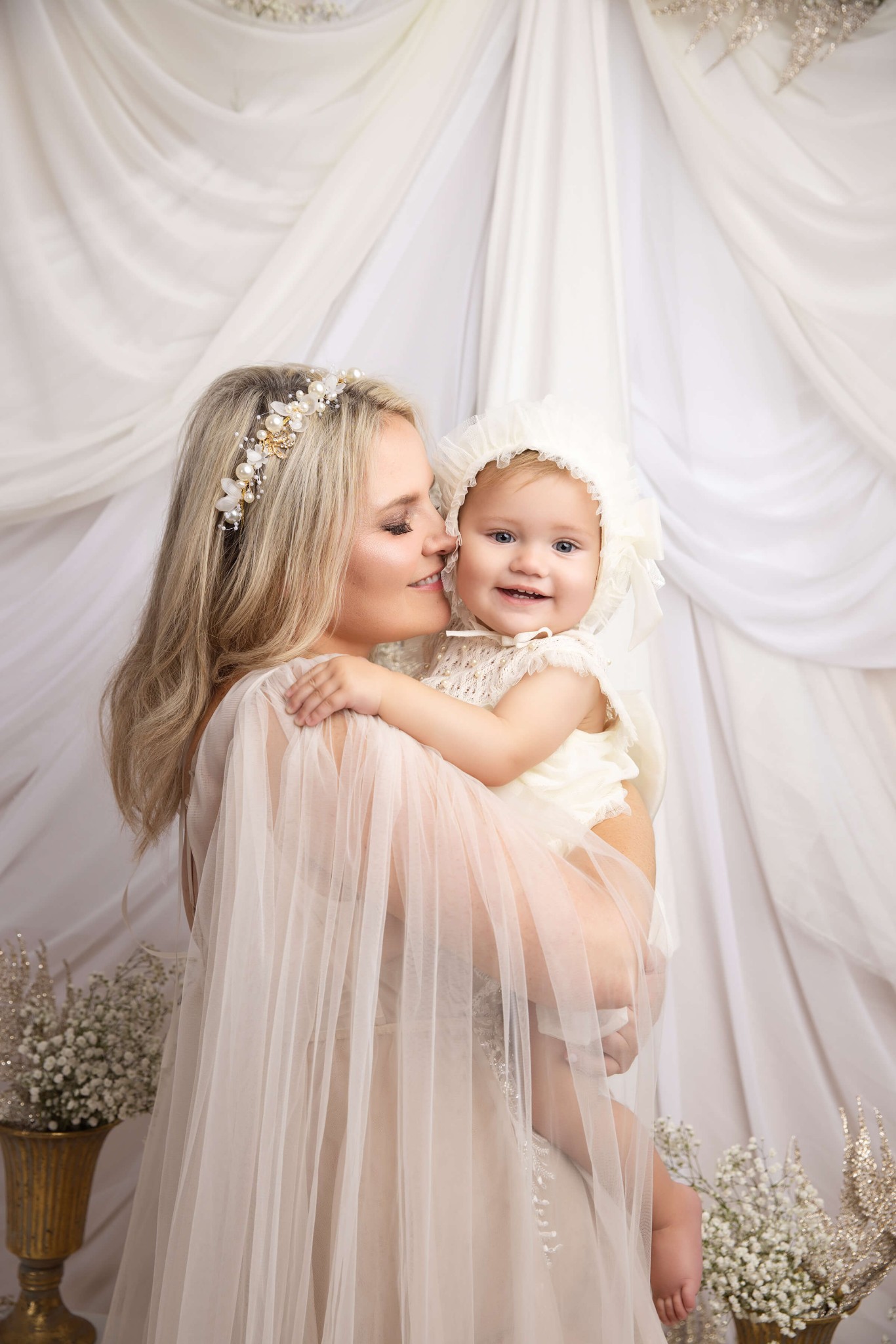 A mom leans in to kiss her infant in her arms both in white in a studio after visiting westfield dance school