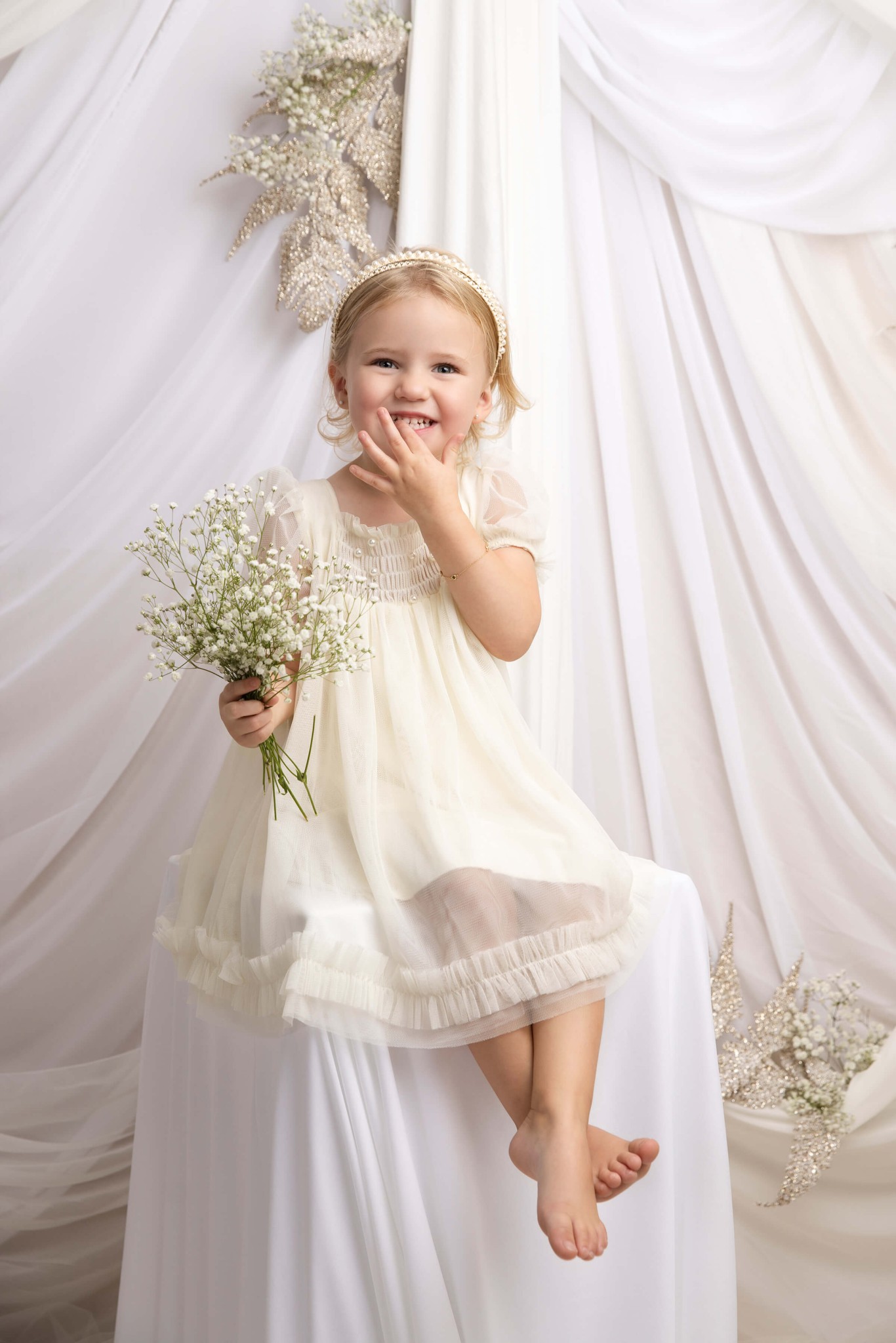 A giggling toddler girl in a white dress holding some wildflowers in a studio after visiting westfield dance school
