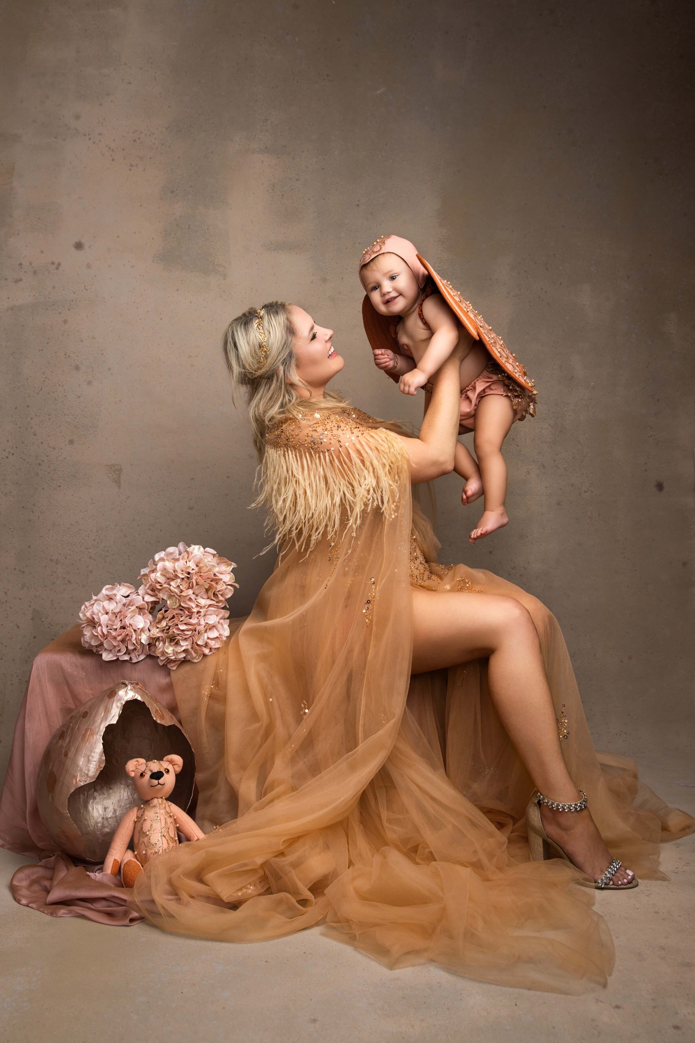 A happy mother in a gold gown lifts her toddler up while sitting in a studio after visiting pediatric dentists in Morristown, NJ
