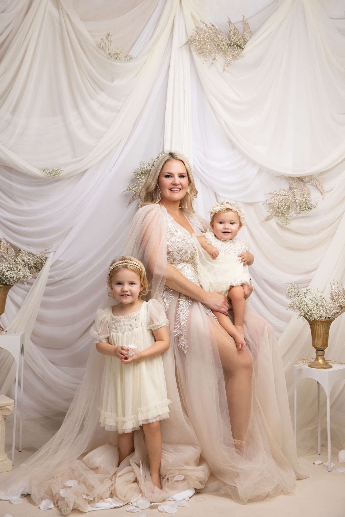 A happy mother in white stands in a studio holding a toddler girl with big sister standing after exploring summer camps in morristown, NJ
