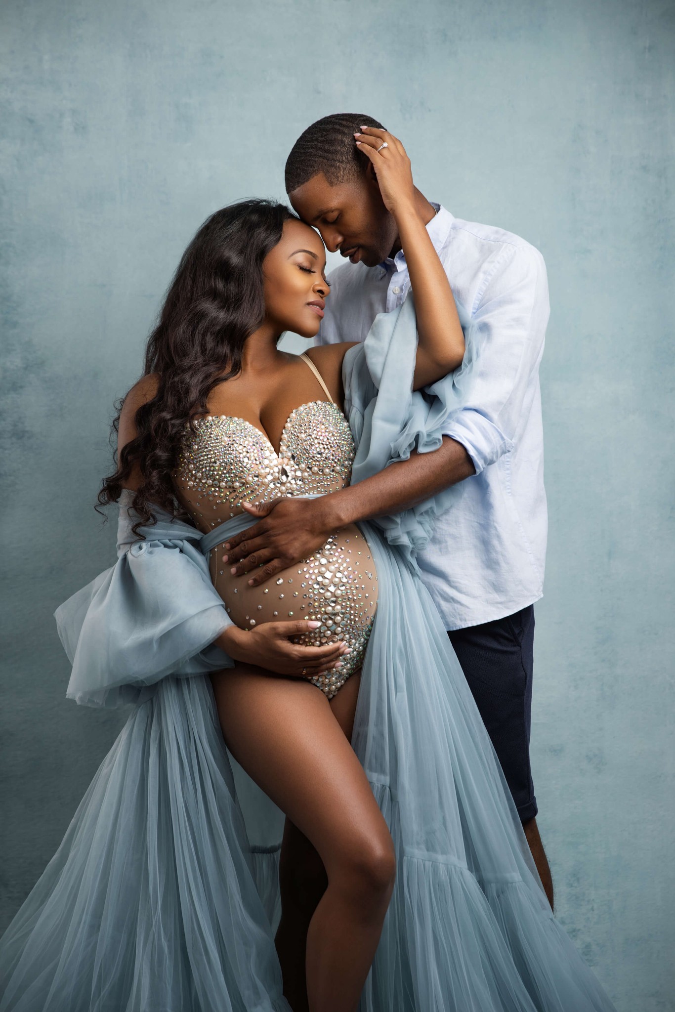 A mom to be in a bodysuit and blue gown snuggles in the arms of her husband in a studio after using care for moms