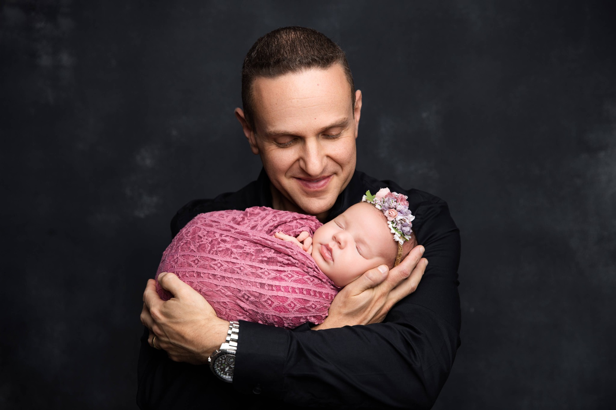 A happy new dad in black cradles his sleeping newborn against his chest in a pink swaddle after finding daycares in summit nj