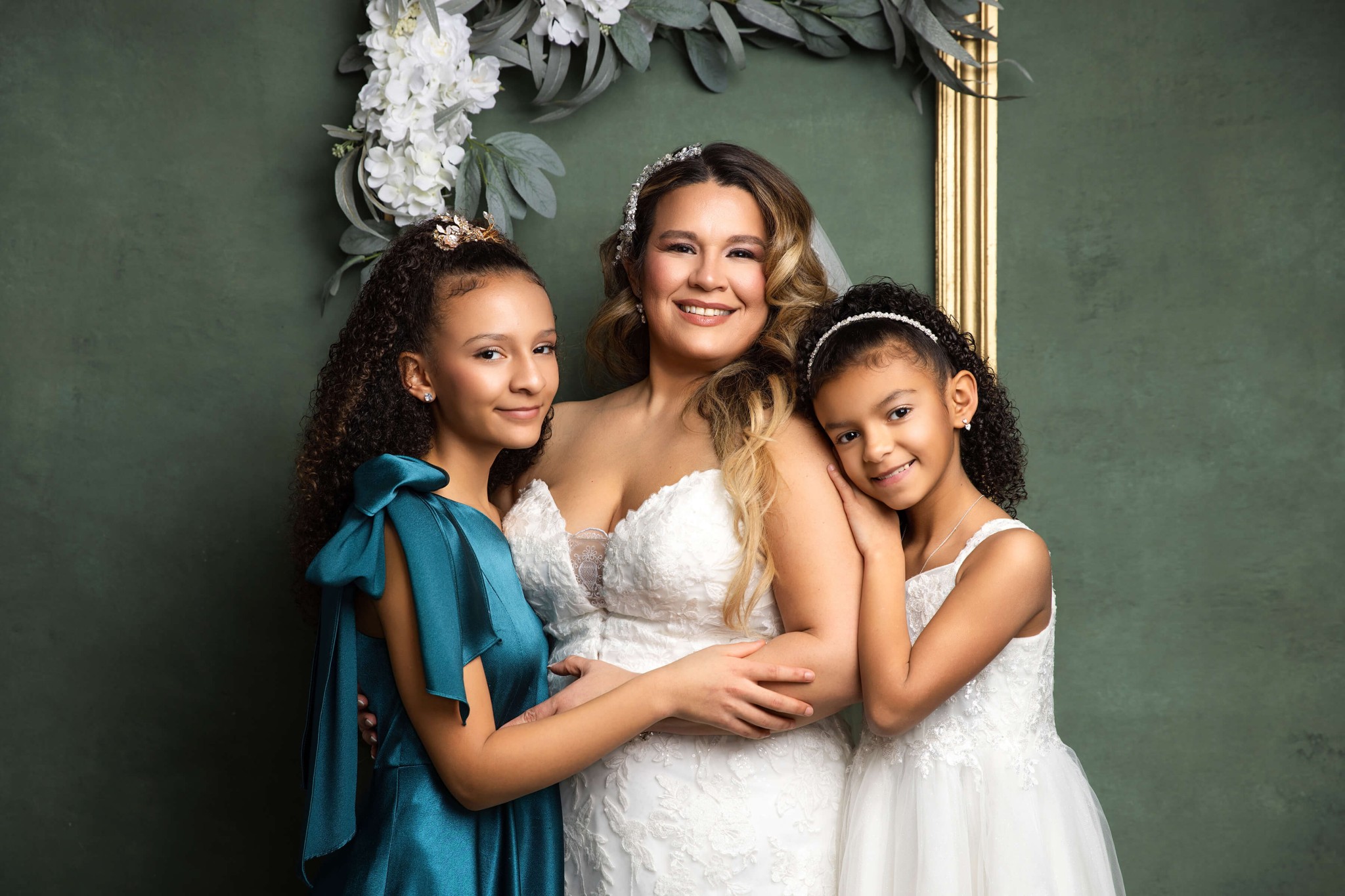 A smiling mother in a white gown stands with her two young daughters in gowns in a studio