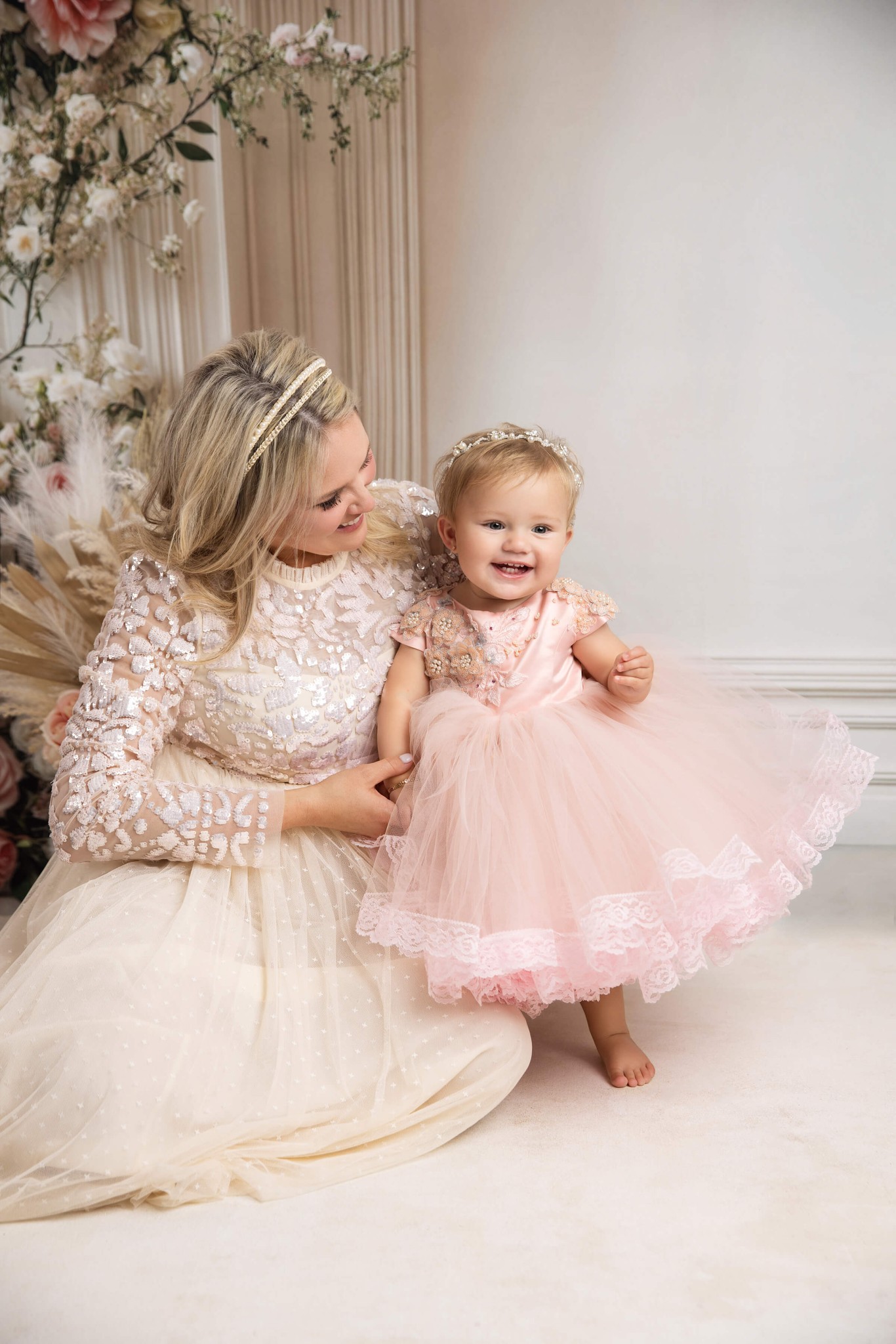 A giggling toddler girl in a pink gown leans against mom in a gold gown on the floor of a studio after finding pediatricians in summit nj