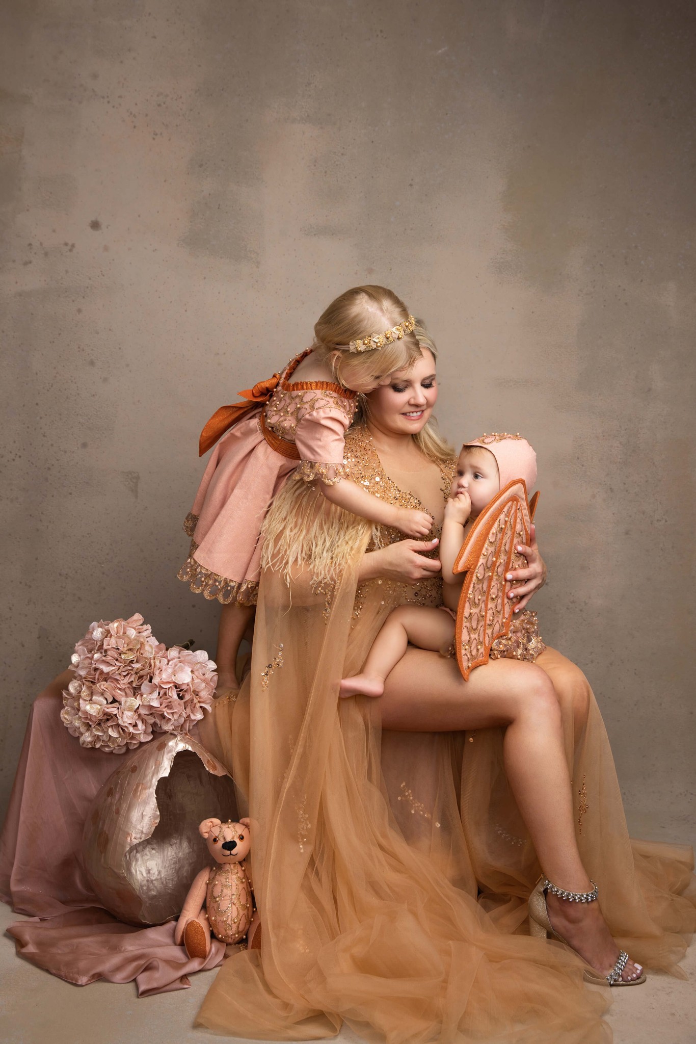 A happy mother sits in a studio in a gold gown with a toddler climbing on her back and one in her lap after finding pediatricians in summit nj