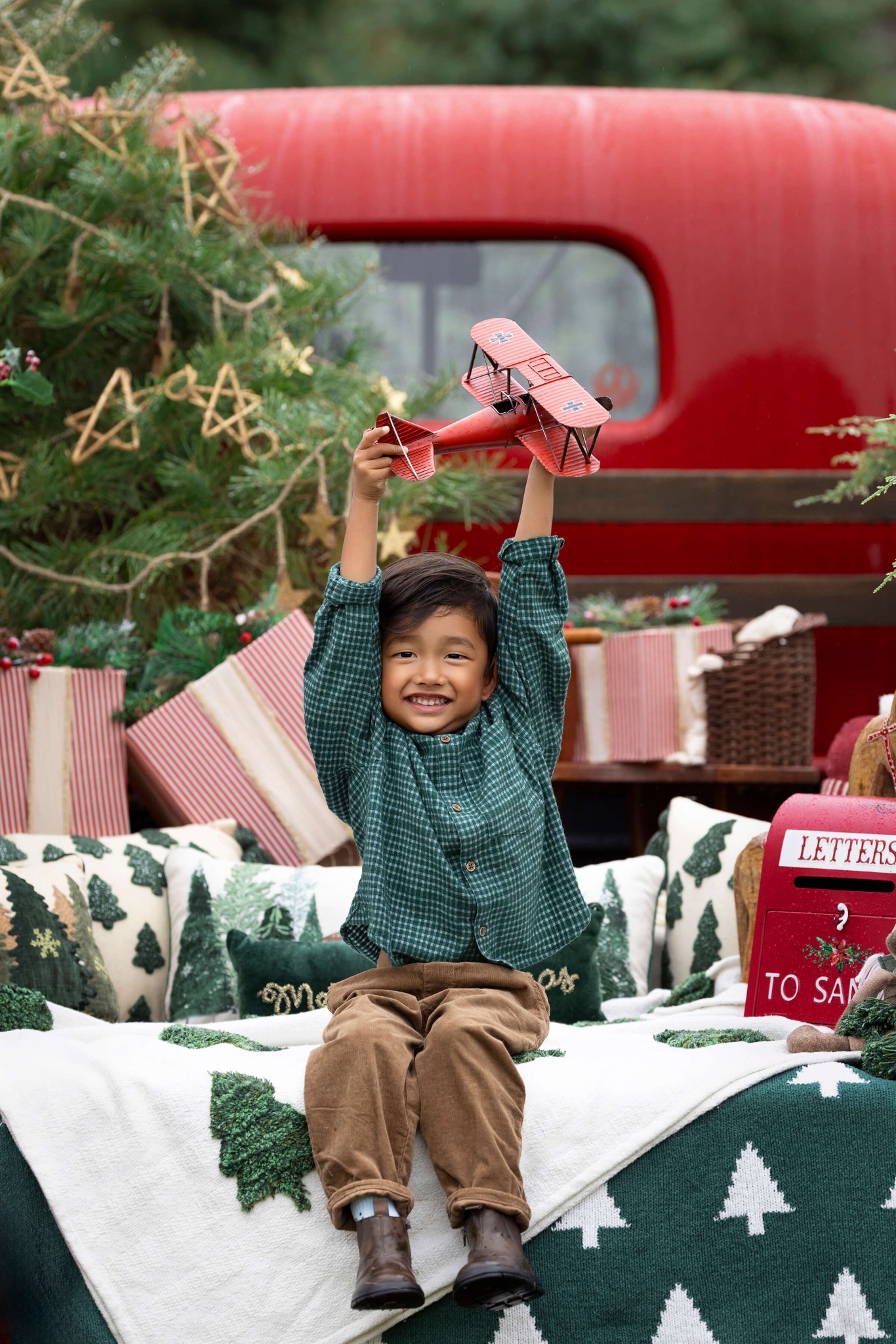 A toddler boy in a green shirt holds up a red toy airplane while sitting in a christmas decorated vintage truck bed after finding preschools in summit nj