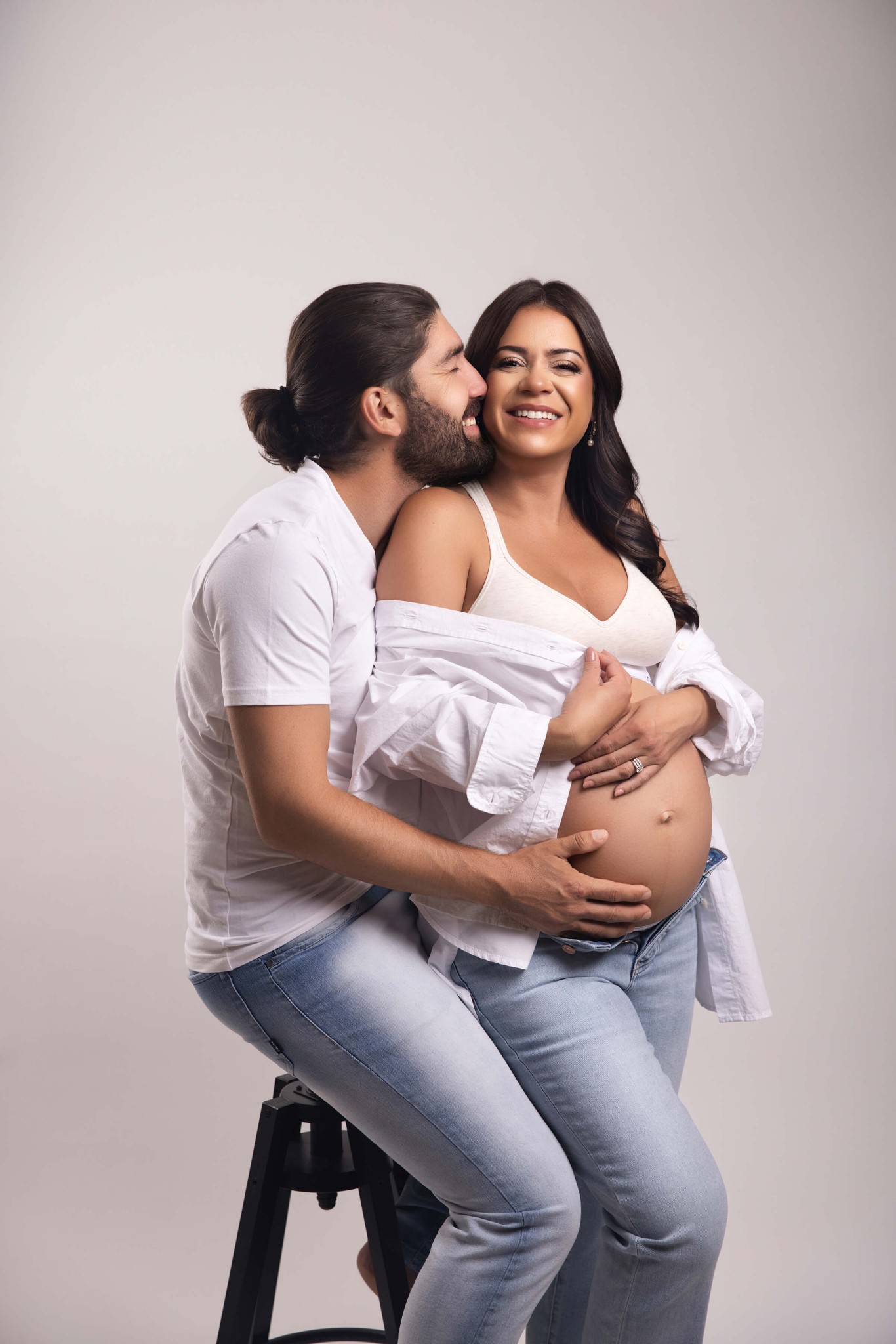 A happy pregnant couple snuggles on a stool in a studio in jeans and white shirts after meeting the women's care source