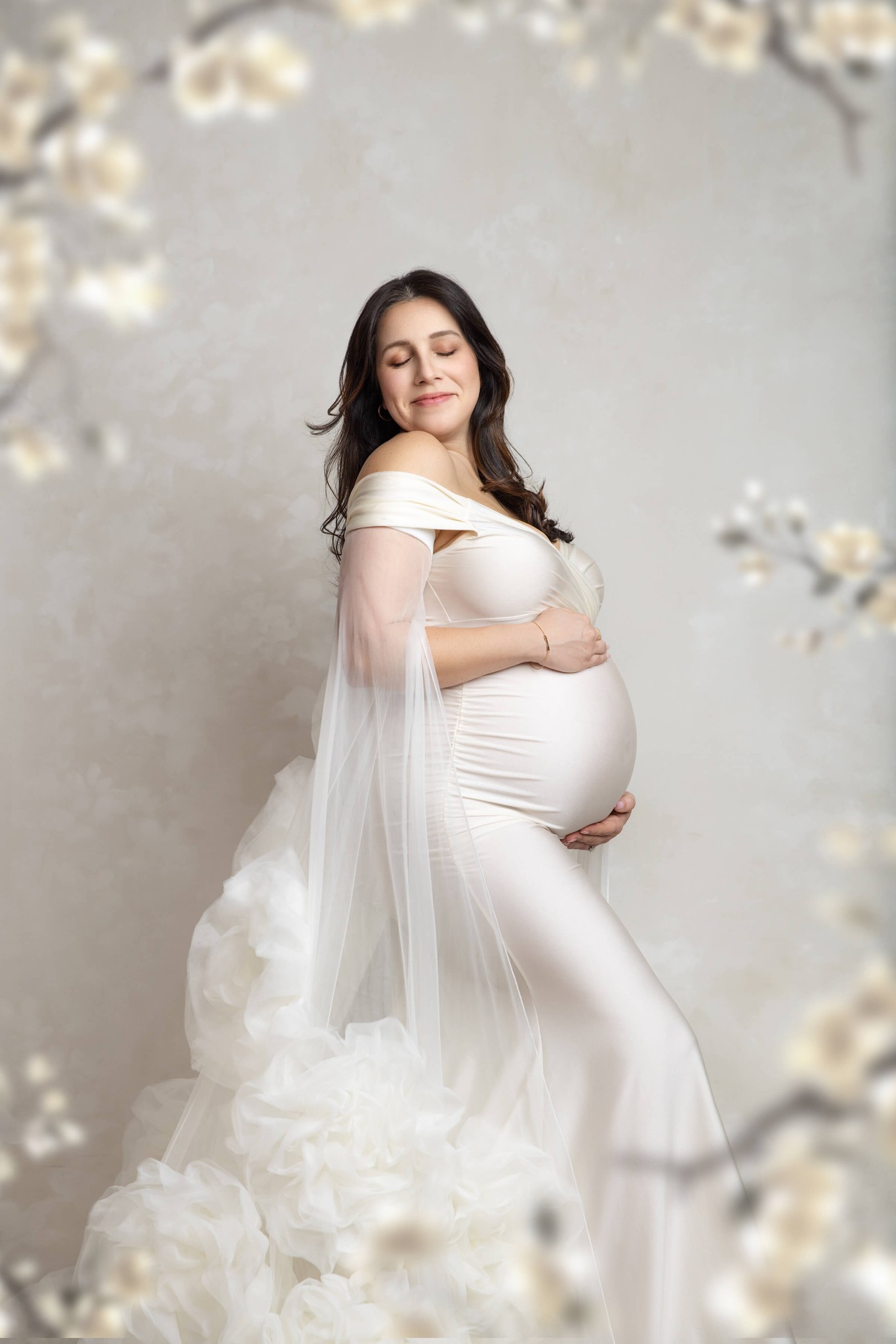 A happy pregnant woman stands in a white maternity gown in a studio in profile with hands on the bump after visiting daycares in millburn nj