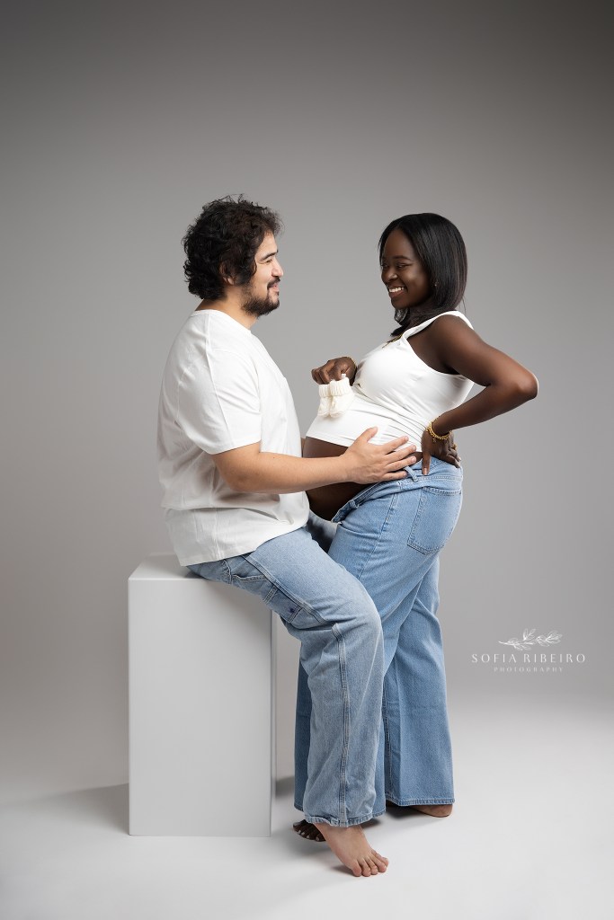 mom and dad share a smile together while holding baby booties against mom's baby bump