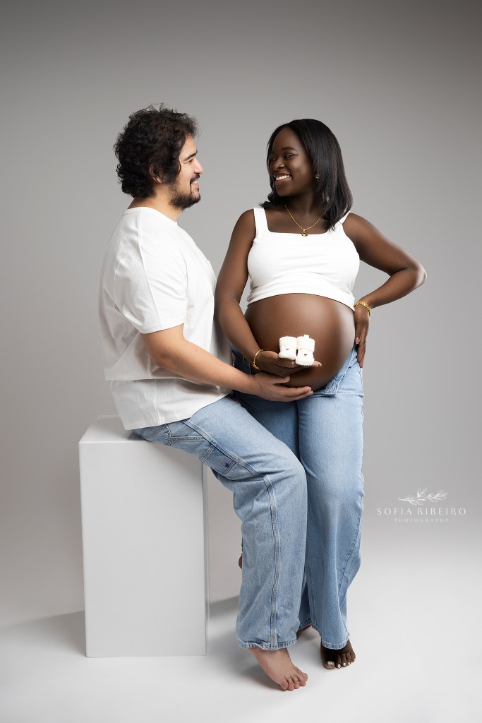 mom and dad share a giggle holding baby booties to the baby bump in matching denim looks in a nj maternity photo studio
