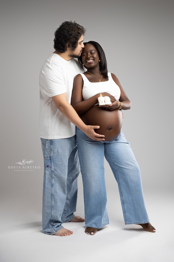 mom and dad share a giggle holding baby booties to the baby bump in matching denim looks in a nj maternity photo studio