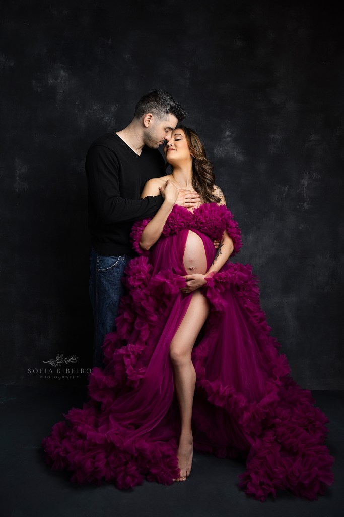 mom and dad to be pose with baby bump in a pink ruffled dress against a black backdrop