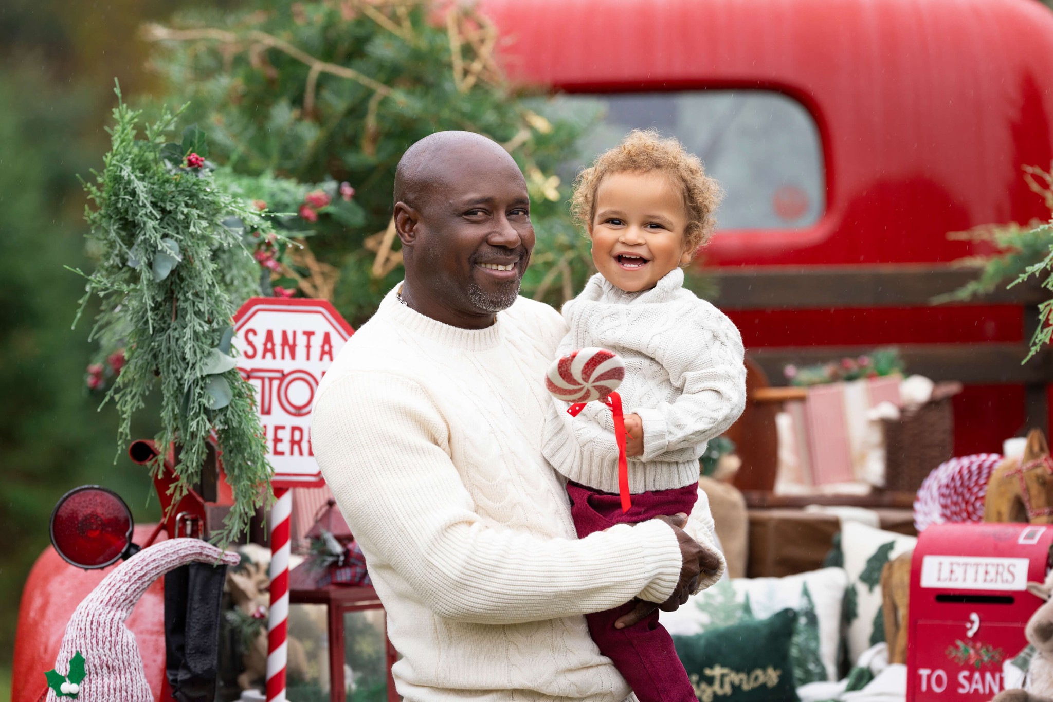 A happy dad in a white sweater stands in front of santa's vintage red pickup truck holding his toddler daughter on his hip after visiting pediatric dentists in morris county nj