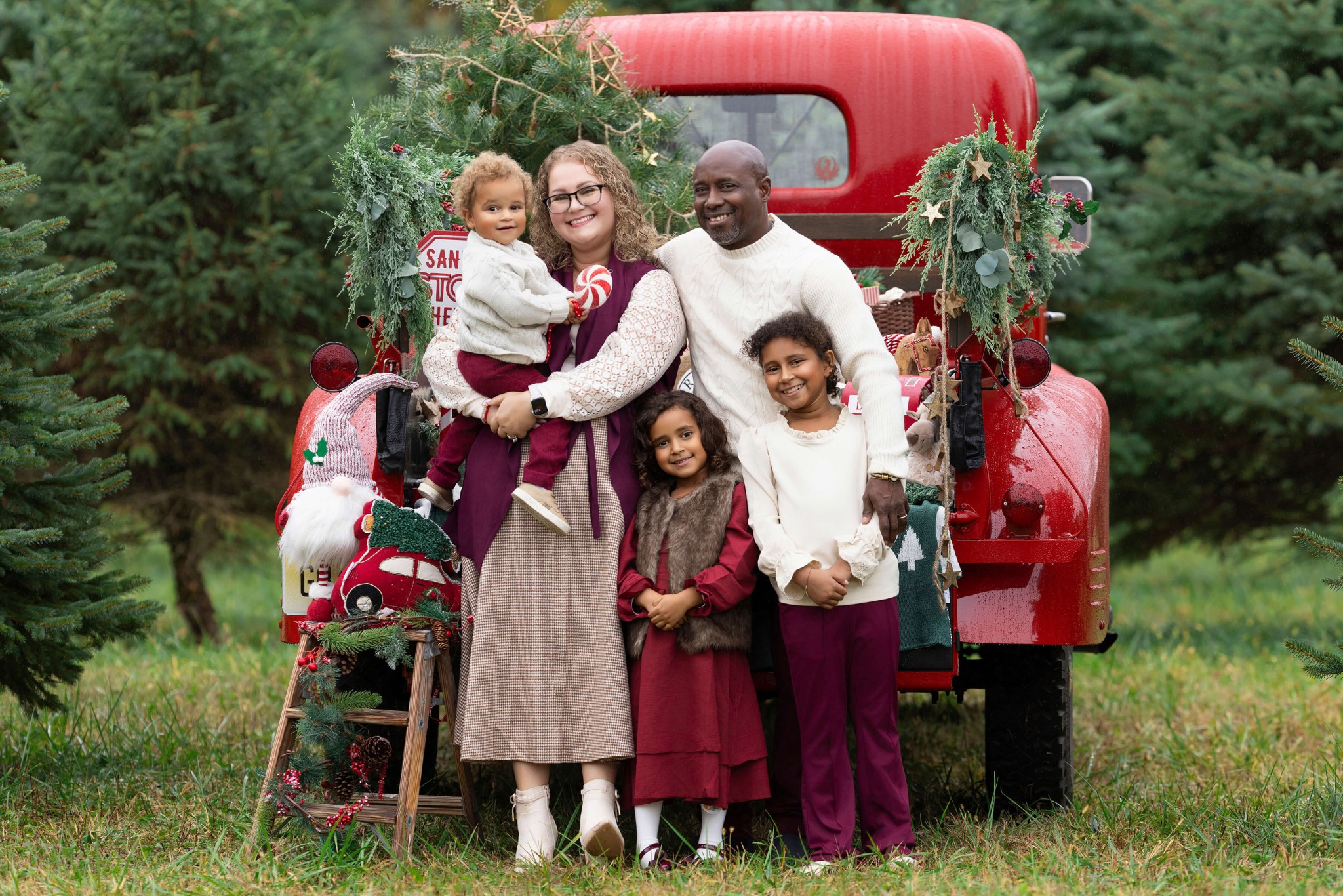 SMiling mom and dad stand against a red vintage truck in sweaters with arms around their three young children after visiting pediatric dentists in morris county nj