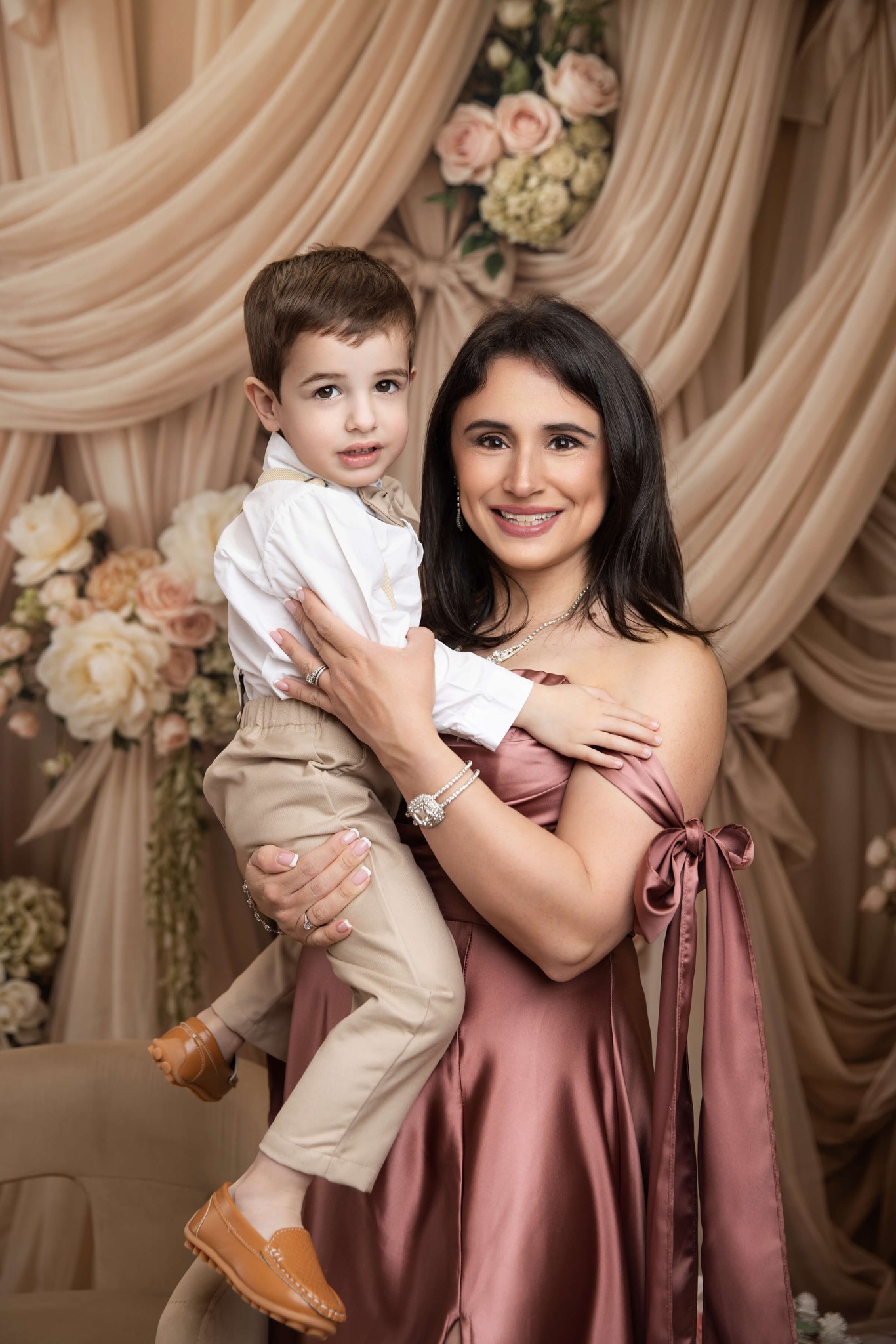 A smiling mother in a pink silk gown holds her toddler son in khakis and white shirt in a studio after visiting preschools in millburn NJ