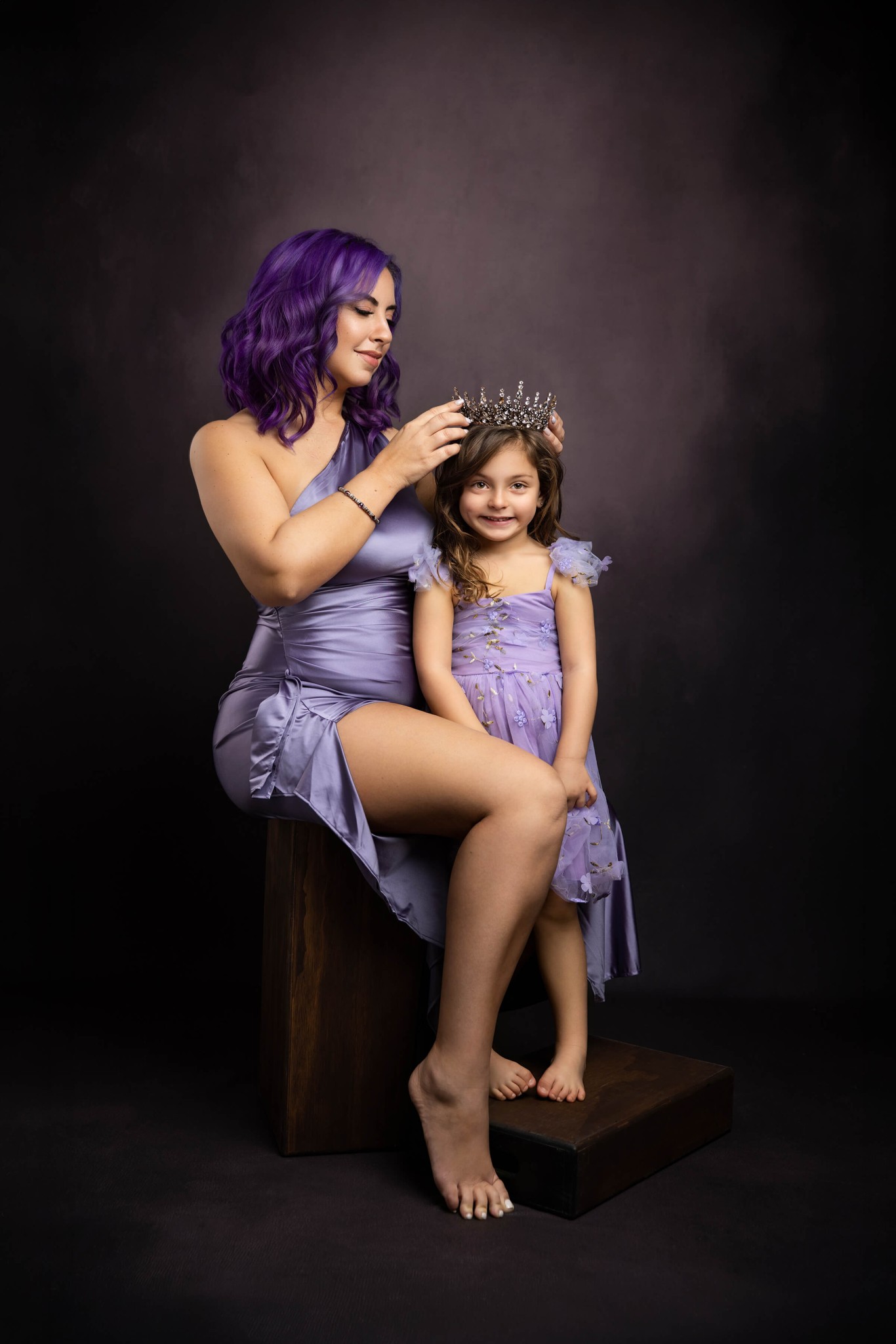 A mother in purple sits on a stool placing a tiara on her daughter's head in matching dress after finding preschools in mendham ,NJ