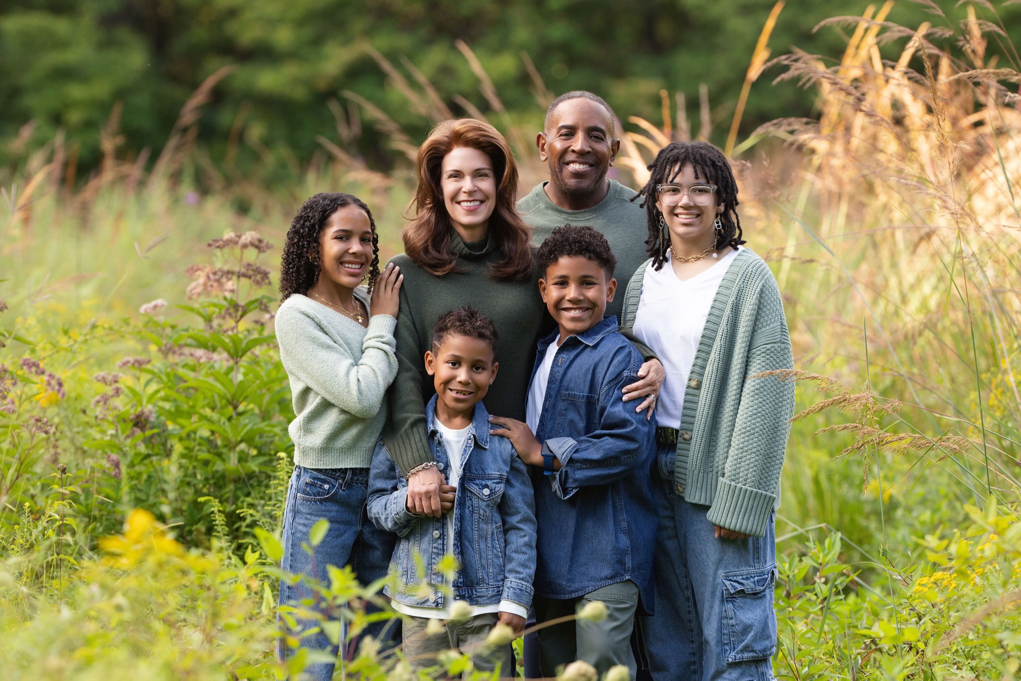 Happy mom and dad stand in a park in tall grasses smiling with their four children in denim and green after enjoying summer camps in Chatham, NJ